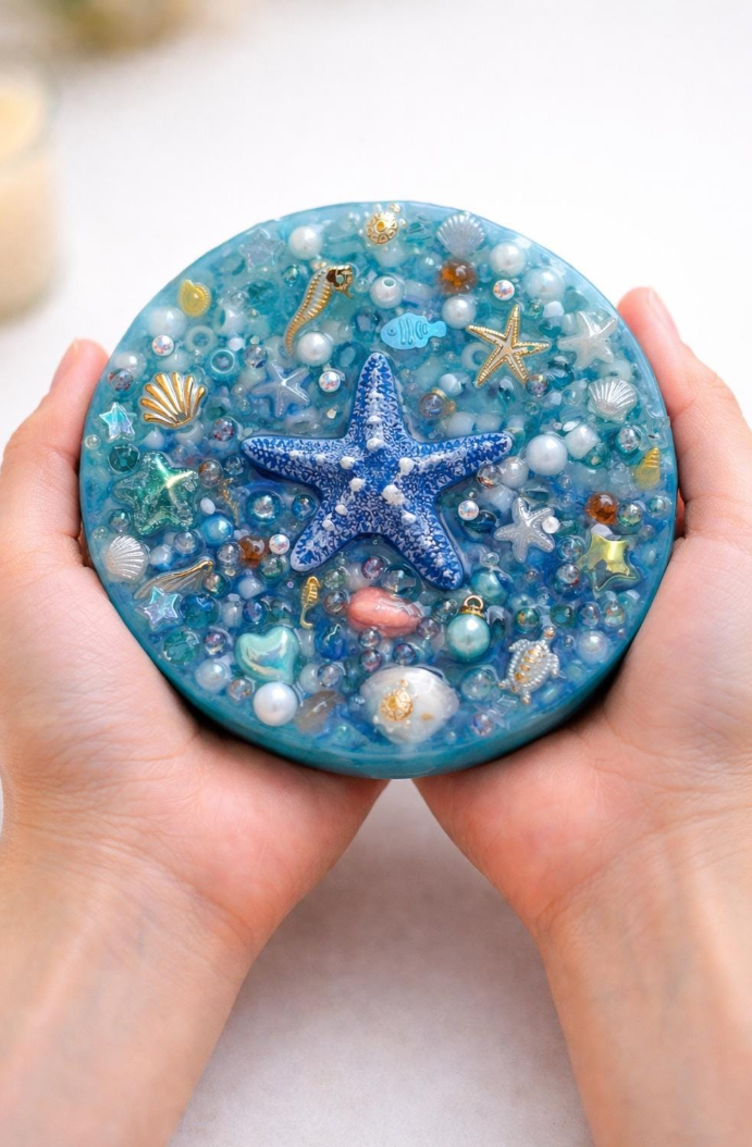 Hand holding a round, ocean-themed resin art piece with blue base and starfish, seashells, and pearls.